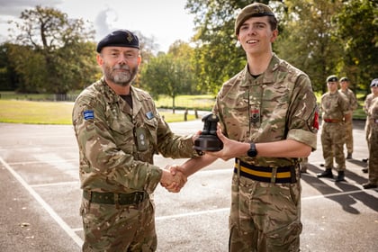 An older officer in camouflage uniform shakes hands with a younger cadet, presenting him with a small trophy.