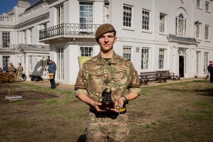 A young soldier in camouflage uniform and a tan beret stands outside a white building, holding a small statuette and a commemorative coin.