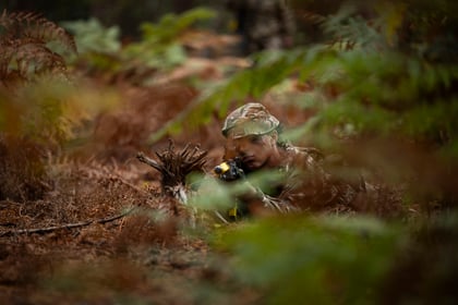 A soldier in camouflage uniform and hat lies prone in a forest, aiming a rifle fitted with a yellow muzzle attachment.