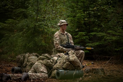 A soldier in camouflage uniform and boonie hat kneels in a forest, holding a rifle with a yellow training attachment, while another soldier lies prone nearby with a rucksack and sleeping mat.