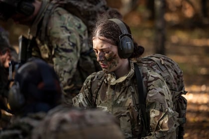 An Army Cadet with camouflage face paint wears a headset and field gear whilst training in a forest.