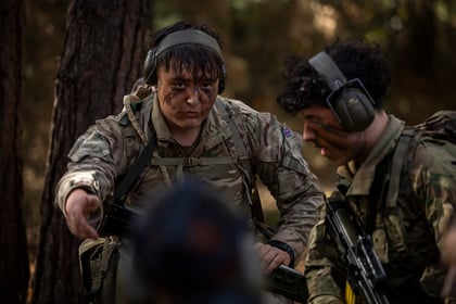 Two soldiers in camouflage uniforms and protective headsets, one with face paint, crouch in a woodland while one gestures towards the camera.