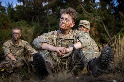 Three Army Cadets in camouflage uniforms and face paint sit in a grassy area.