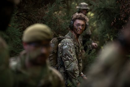 A cadet with camo cream and a headset looks back whilst patrolling through a forest with other cadets.