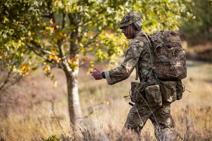 A cadet in a camouflage uniform and a large rucksack walks through a field, gesturing with one hand.