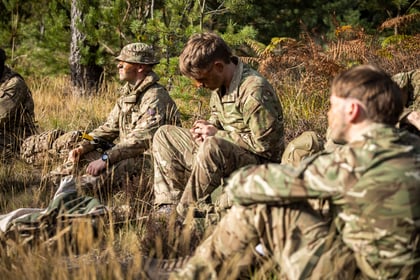 Three cadets in camouflage uniforms are resting in a grassy, wooded area.