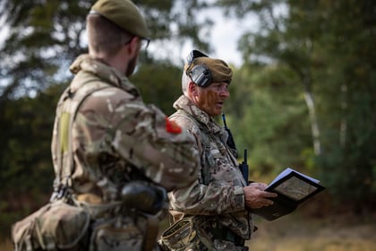 Two soldiers in camouflage uniforms and berets discuss a map in a wooded area.