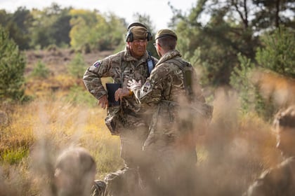 Two Army Officers in camouflage uniforms consult a notebook whilst standing in a grassy field.