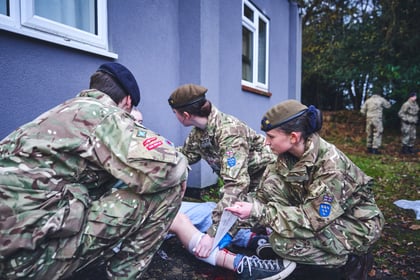 Three uniformed military cadets provide first aid to a casualty's injured leg outside a building.