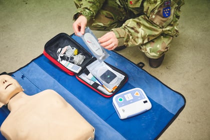 A person in camouflage uniform prepares an AED trainer kit and electrode pads next to a CPR training manikin.