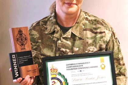 A woman in a military uniform holding a wooden award trophy and a framed certificate for the Welsh Ambulance Services Volunteer Conference Awards.