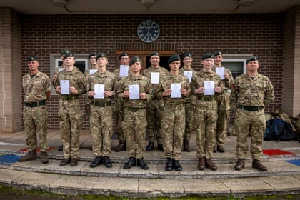 Twelve soldiers in camouflage uniforms stand in two rows in front of a brick building, each holding a white certificate.