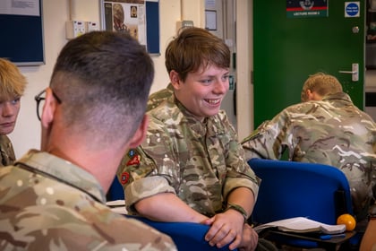Three people in military camouflage uniforms sitting in a classroom, with one smiling person in the centre looking towards a person whose back is to the camera.