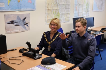 A woman in a navy dress and mayoral chain sits beside a young man in a flight suit who gestures while using a flight simulator joystick.
