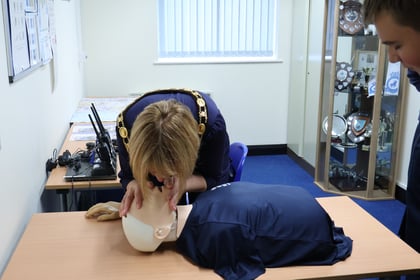A woman wearing a ceremonial chain of office performs CPR on a medical training manikin while a person looks on.