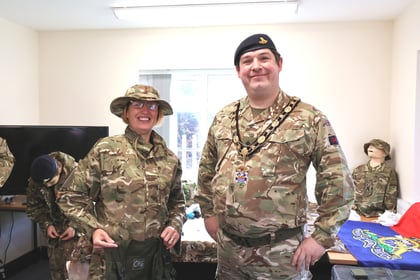 Two people in camouflage uniforms, one wearing a boonie hat and the other a beret and a formal military chain of office, stand smiling in a room.