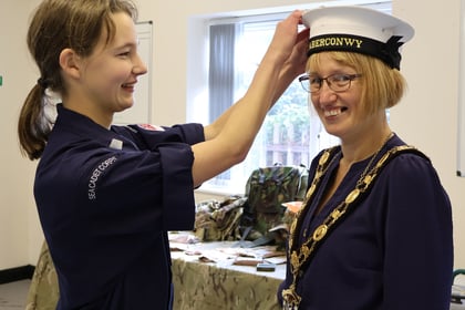 A Sea Cadet places a white cap labelled ABERCONWY on the head of a smiling woman wearing a ceremonial chain of office.