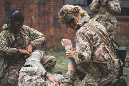 Two soldiers in camouflage uniforms provide medical aid to a third soldier lying on the ground.