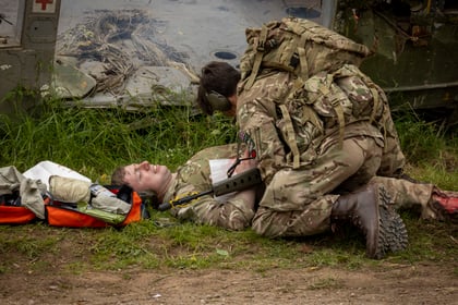 A soldier in camouflage gear kneels over an injured soldier lying on the grass to provide medical aid.