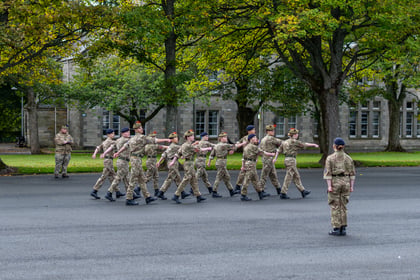 A group of soldiers in camouflage uniforms marching in formation on a tarmac parade ground whilst a soldier in uniform stands in the foreground observing them.