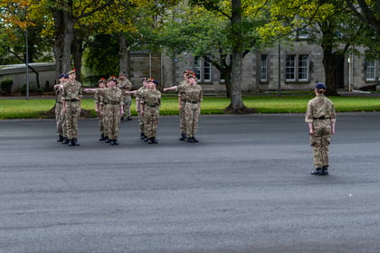 A group of cadets in camouflage uniforms perform drills on a paved parade ground whilst facing an instructor.