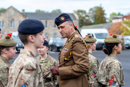 A British Army officer in a dress uniform and beret speaks to a group of cadets in camouflage uniforms.