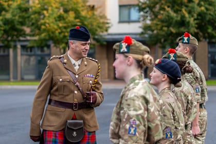 An officer in a brown uniform and kilt speaks to a line of four soldiers in camouflage uniforms and berets.