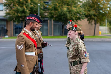 A British soldier in a formal tan uniform and a red-chequered glengarry cap smiles at a female soldier in camouflage uniform during a military inspection.