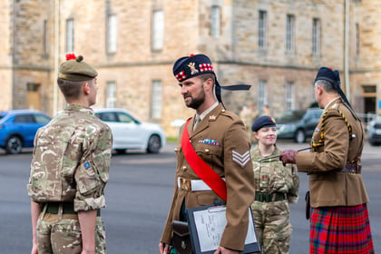 Three British military personnel in uniform stand outdoors, with one officer in a kilt and a Tam o' Shanter cap speaking to a younger soldier.
