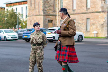 A woman in camouflage military fatigues and a man in a formal brown uniform with a tartan kilt standing in a car park.