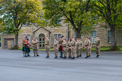 A group of soldiers in camouflage uniforms and two bagpipers in kilts stand in formation in front of a stone building.