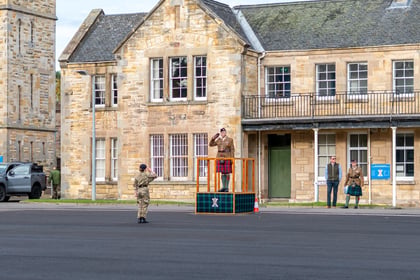 A soldier in a kilt salutes whilst standing on a small wooden platform in front of a stone building, as another soldier in camouflage salutes from the ground nearby.