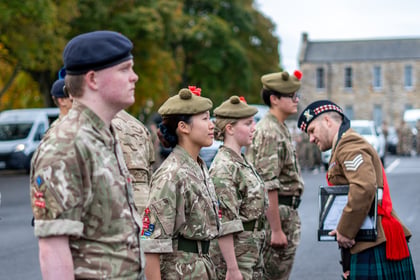 A uniformed soldier in a kilt hands a folder to a line of standing cadets wearing camouflage uniforms and berets.