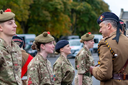 A military officer in a dark beret speaks to a line of soldiers in camouflage uniforms and tan berets.