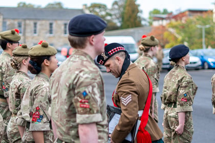 A sergeant in a brown ceremonial uniform with a red sash stands among a group of soldiers in camouflage uniforms during an outdoor parade.