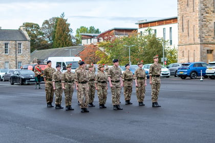A group of ten cadets in camouflage uniforms standing in formation on an asphalt parade ground, with a lone bagpiper standing in the background.
