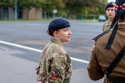A young woman in military camouflage uniform and dark beret stands outdoors, with two other service members visible in the foreground and background.