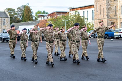 A group of cadets in camouflage uniforms marching and saluting in a paved car park.