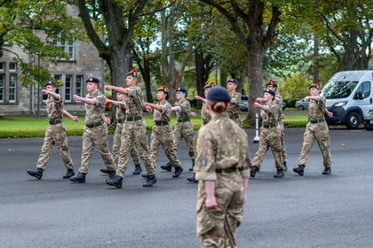 A group of army cadets in camouflage uniforms march in formation on a tarmacked area, led by an individual in the foreground.
