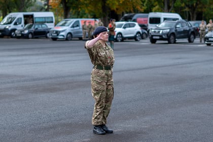 A soldier in camouflage uniform and a beret stands to attention and salutes in a tarmacked car park with several vehicles in the background.
