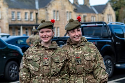 Two smiling Army cadets in camouflage uniforms and tam o’ shanter hats standing outdoors.