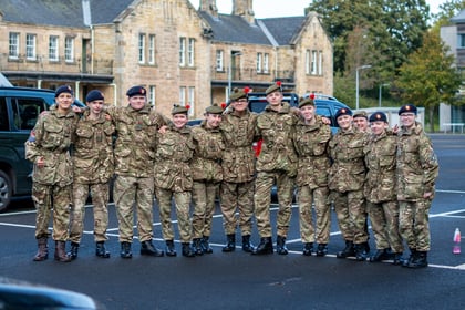 A group of eleven young people in military uniforms and berets standing in a row with their arms round each other's shoulders in a car park.