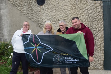 Four people stand outdoors holding a green and black banner for a Normandy Battlefield Tour 2026 that reads Supported by Armed Forces Cadet Charitable Trust UK.