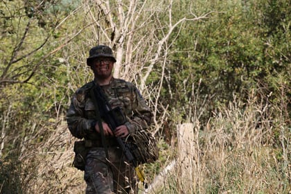 A person in camouflage military uniform and helmet stands in tall grass with a rifle, surrounded by trees and dense vegetation, participating in the intense 51 Brigade Military Skills Competition.