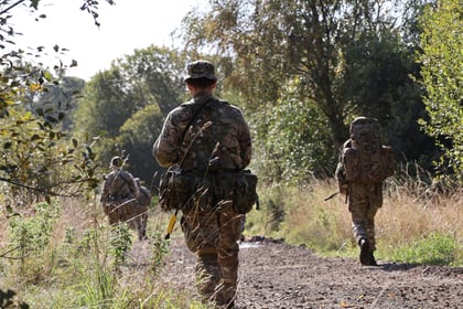 Three soldiers in camouflage uniforms and kit walk along a dirt path through a sunlit, wooded area, with green foliage and trees surrounding them during the 51 Brigade Military Skills Competition.
