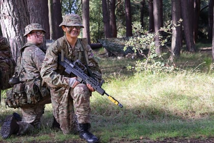 Two soldiers in camouflage uniforms and hats are kneeling in a sunlit forest clearing during the 51 Brigade Military Skills Competition. One soldier is smiling and holding a rifle, whilst the other looks ahead. Trees and greenery surround them.
