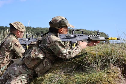 Two soldiers in camouflage uniforms lie prone on grassy terrain, aiming rifles with yellow attachments, likely during a training exercise as part of the 51 Brigade Military Skills Competition. The sky is clear and there are trees in the background.