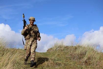 A soldier in camouflage uniform, helmet, and glasses walks through grassy dunes carrying a rifle under a bright blue sky with some clouds, participating in the 51 Brigade Military Skills Competition.