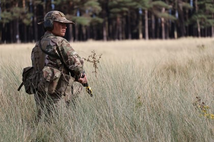 A soldier in camouflage gear and helmet stands in tall grass, looking to the side, with a dense forest in the background—an intense moment captured during the 51 Brigade Military Skills Competition.