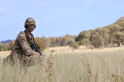 A soldier in camouflage uniform and helmet crouches with a rifle in tall grass, with trees and a blue sky in the background, taking part in the 51 Brigade Military Skills Competition.